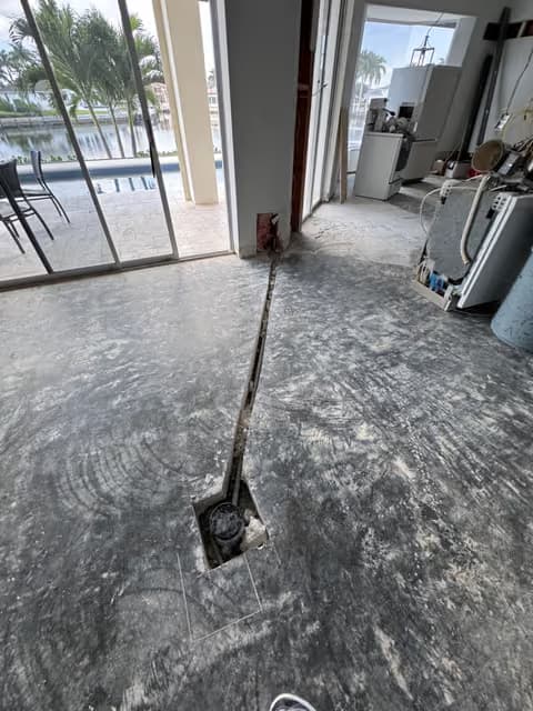 Corte de acceso de plomería en un piso de concreto que lleva a una válvula de cierre, con una terraza de piscina visible a través de la puerta corrediza